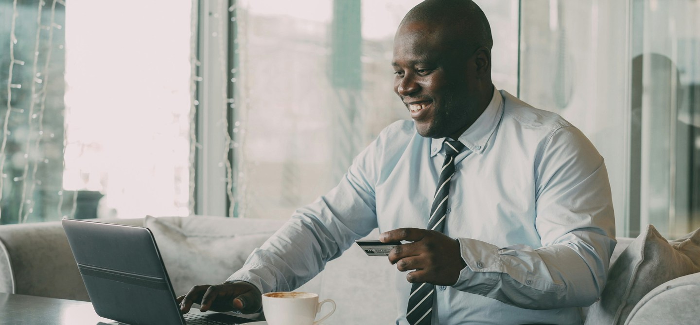 A smiling professional sits on a sofa by a large window, using a laptop while holding a credit card, with a cup of coffee on the table, suggesting an online financial transaction or digital banking activity.
