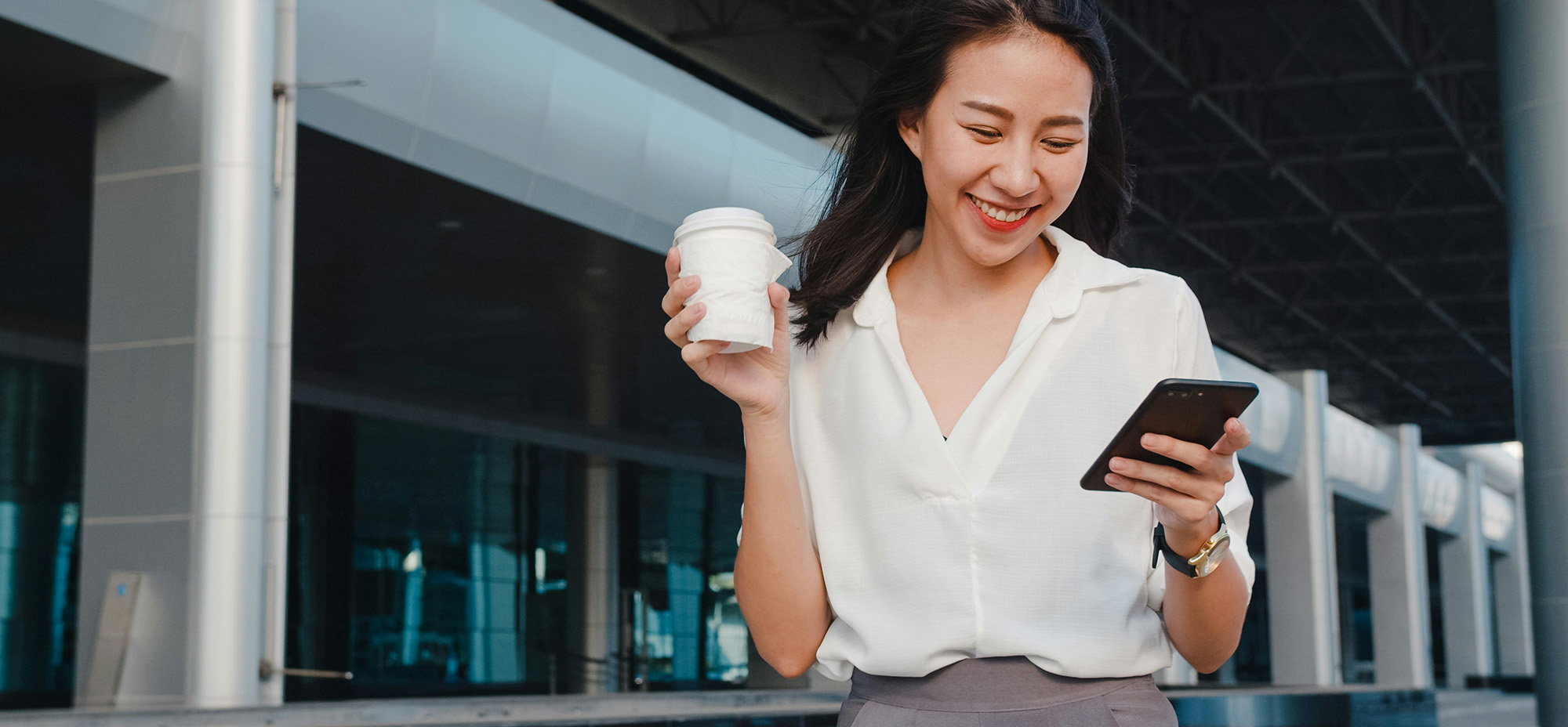Smiling woman outdoors holding a coffee and looking at her smartphone in front of a modern building, illustrating a personalized digital interaction powered by mobile technology.