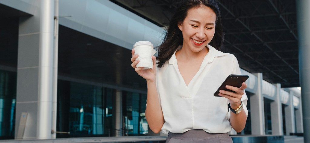 Smiling woman outdoors holding a coffee and looking at her smartphone in front of a modern building, illustrating a personalized digital interaction powered by mobile technology.