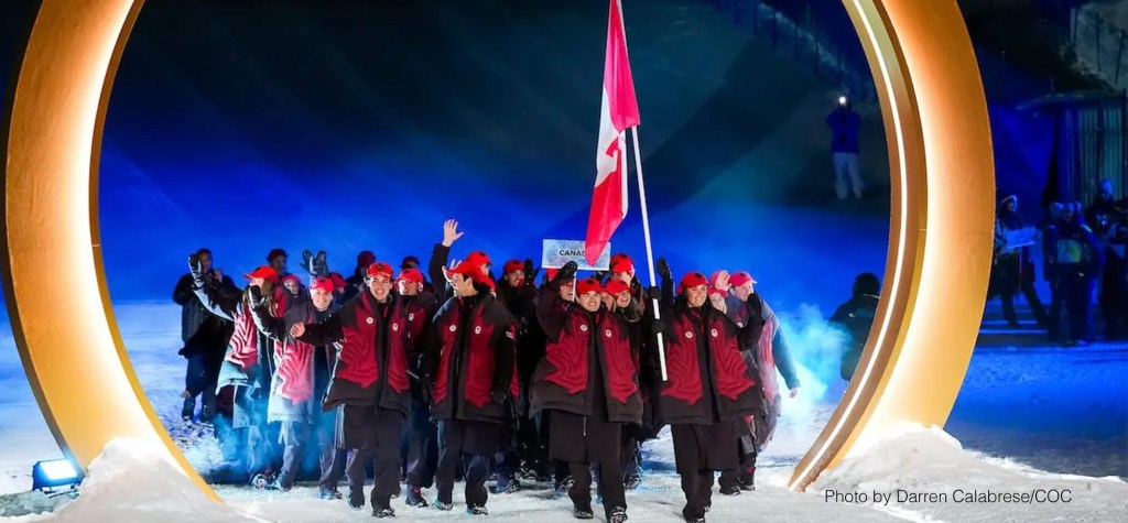 Athletes from Team Canada march into the stadium during a Winter Olympic Games opening ceremony, wearing red jackets and caps while carrying the Canadian flag.