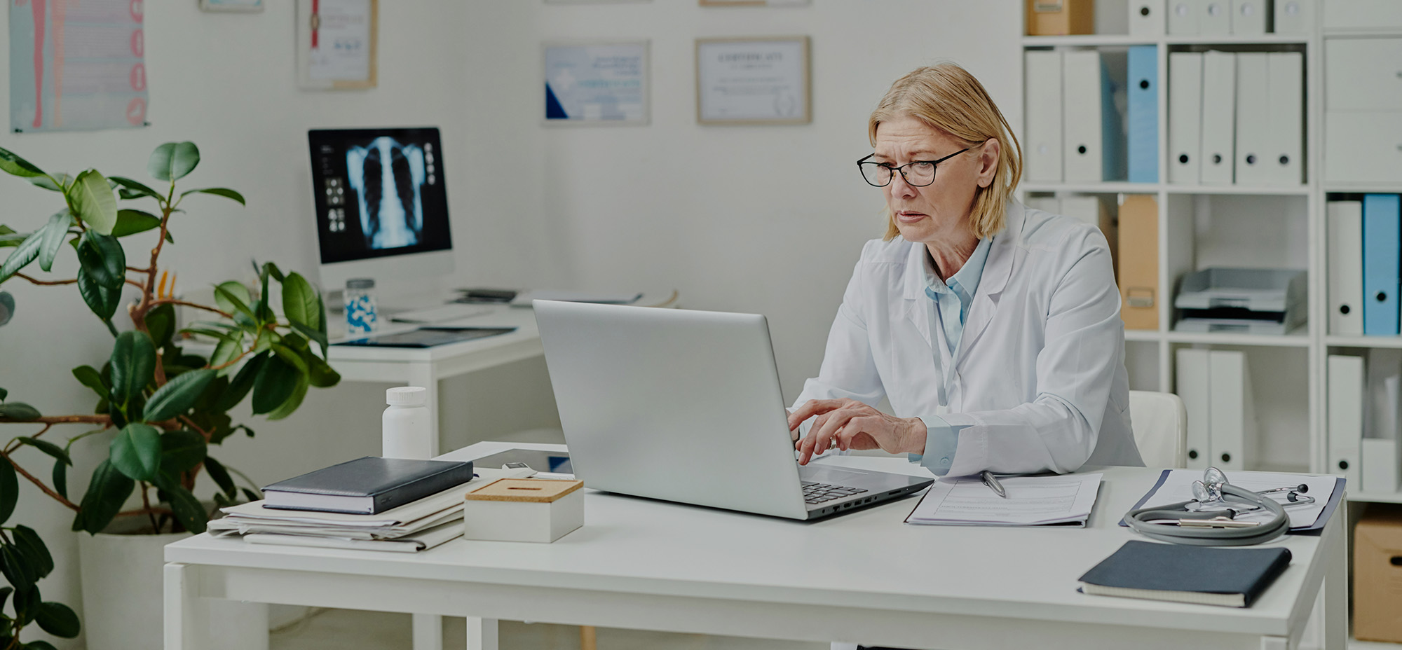 Doctor in a medical office working on a laptop at a desk with medical documents and a stethoscope, with an X-ray image visible on a monitor in the background.