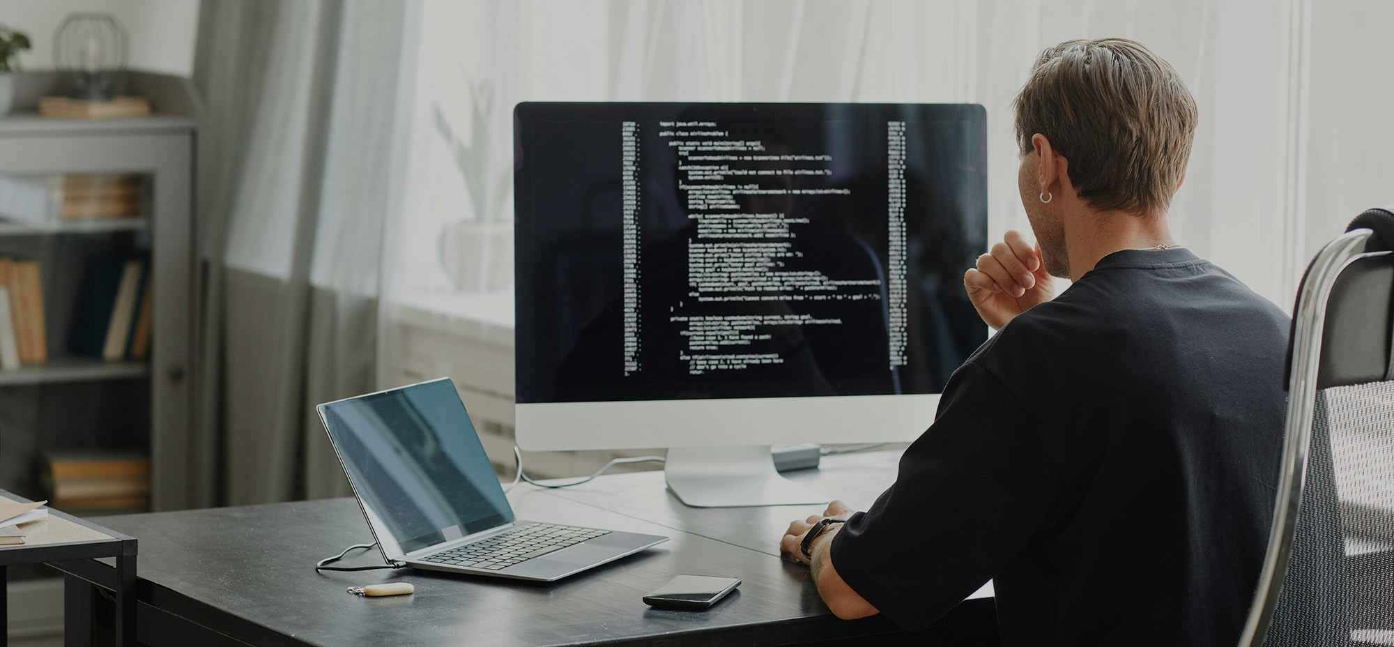 Person working at a desk with a laptop and monitor displaying code, focused on software development in a home office setting.