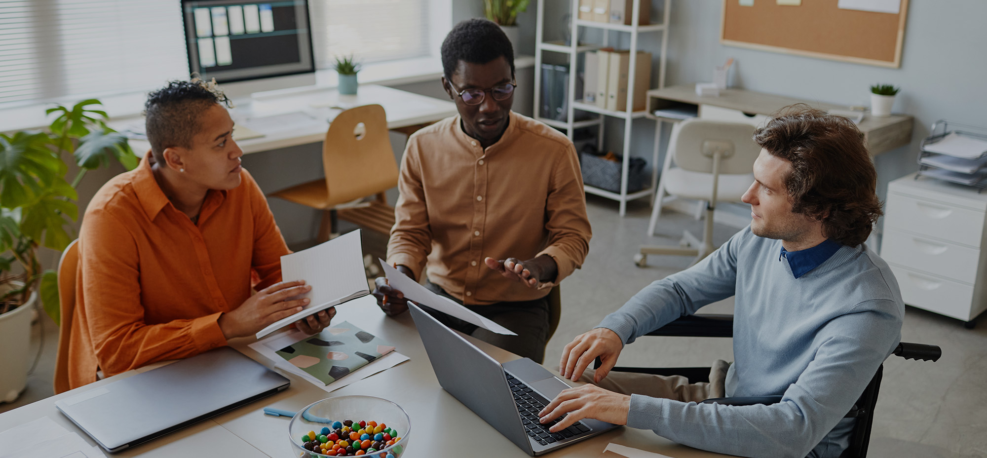 Three colleagues sitting together in an office, discussing documents and working on a laptop.