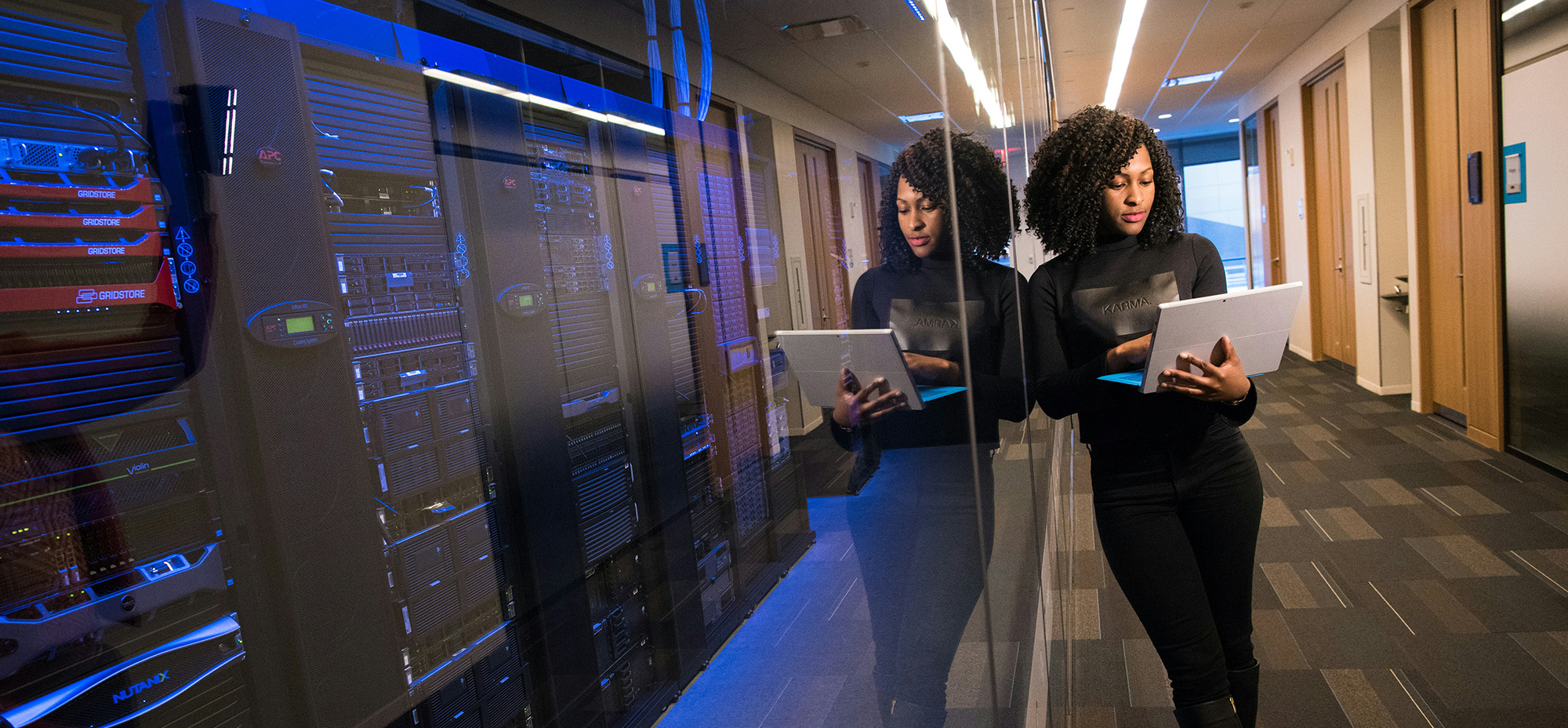 Woman walking through a modern server room while holding a tablet, with rows of enterprise server racks illuminated in blue lighting behind glass panels.