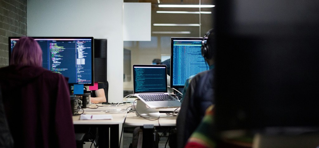 Developers working in a modern office environment with multiple monitors displaying code and system dashboards, representing software development and technical operations.
