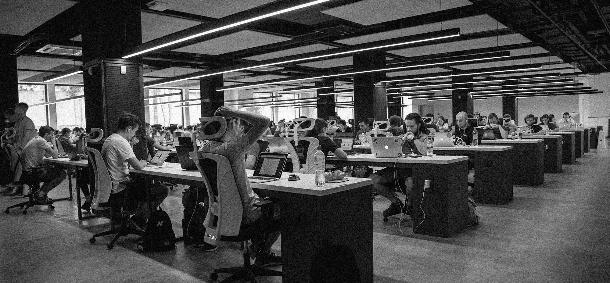 Black-and-white photo of a large contributor or developer workspace filled with people working on laptops at long shared tables under modern ceiling lights.