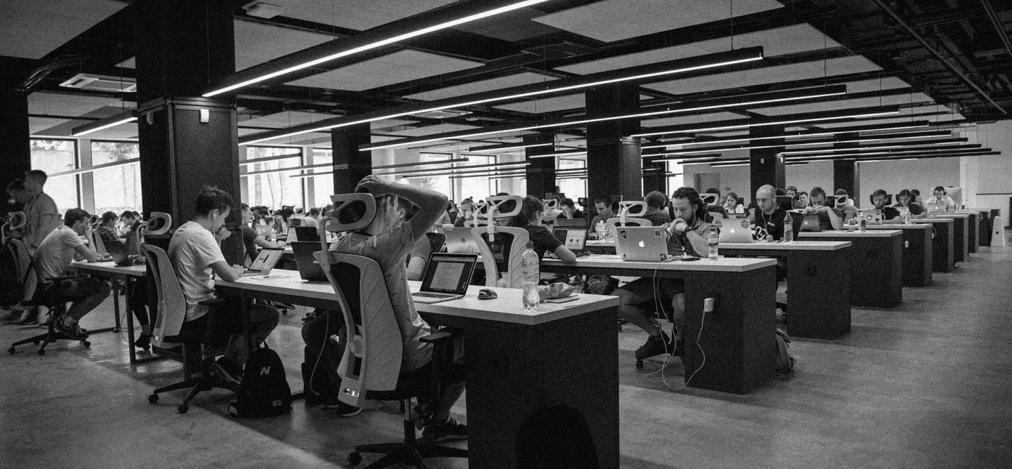Black-and-white photo of a large contributor or developer workspace filled with people working on laptops at long shared tables under modern ceiling lights.
