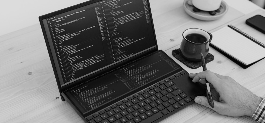 Black-and-white photo of a developer working at a dual-screen laptop displaying code. A hand holding a pen rests near the keyboard, with a coffee cup, notebook, and smartphone on a wooden desk, representing software development, programming, and digital workflows.