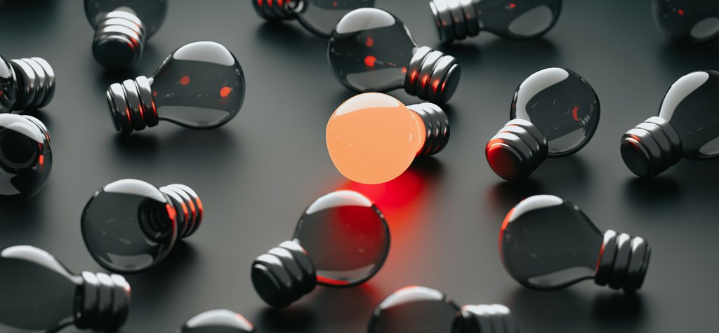 One illuminated lightbulb glowing among several dark, unlit bulbs on a black surface, representing innovation, insight, or identifying opportunities for AI transformation.