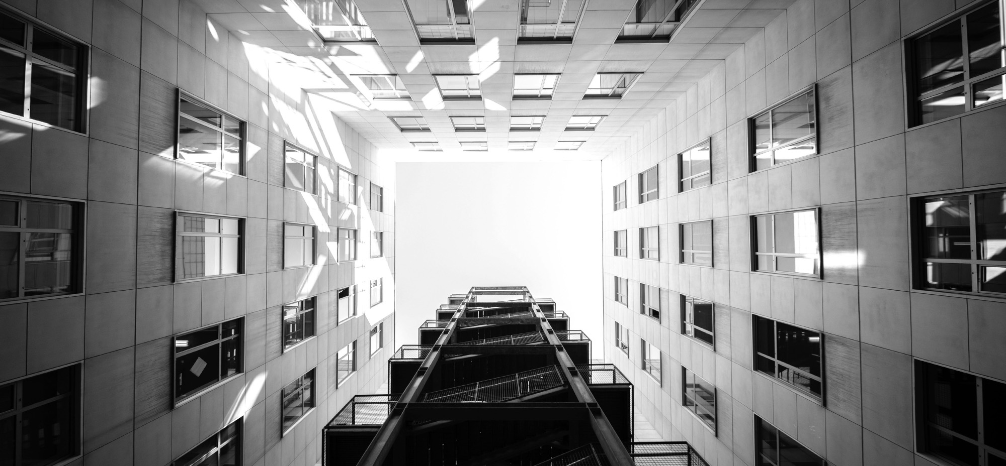 Black-and-white upward view between two modern buildings toward a central exterior staircase and bright sky.
