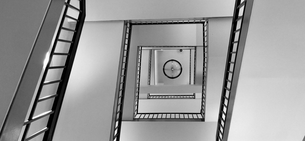 Black-and-white view looking upward through a geometric stairwell with repeating square railings.