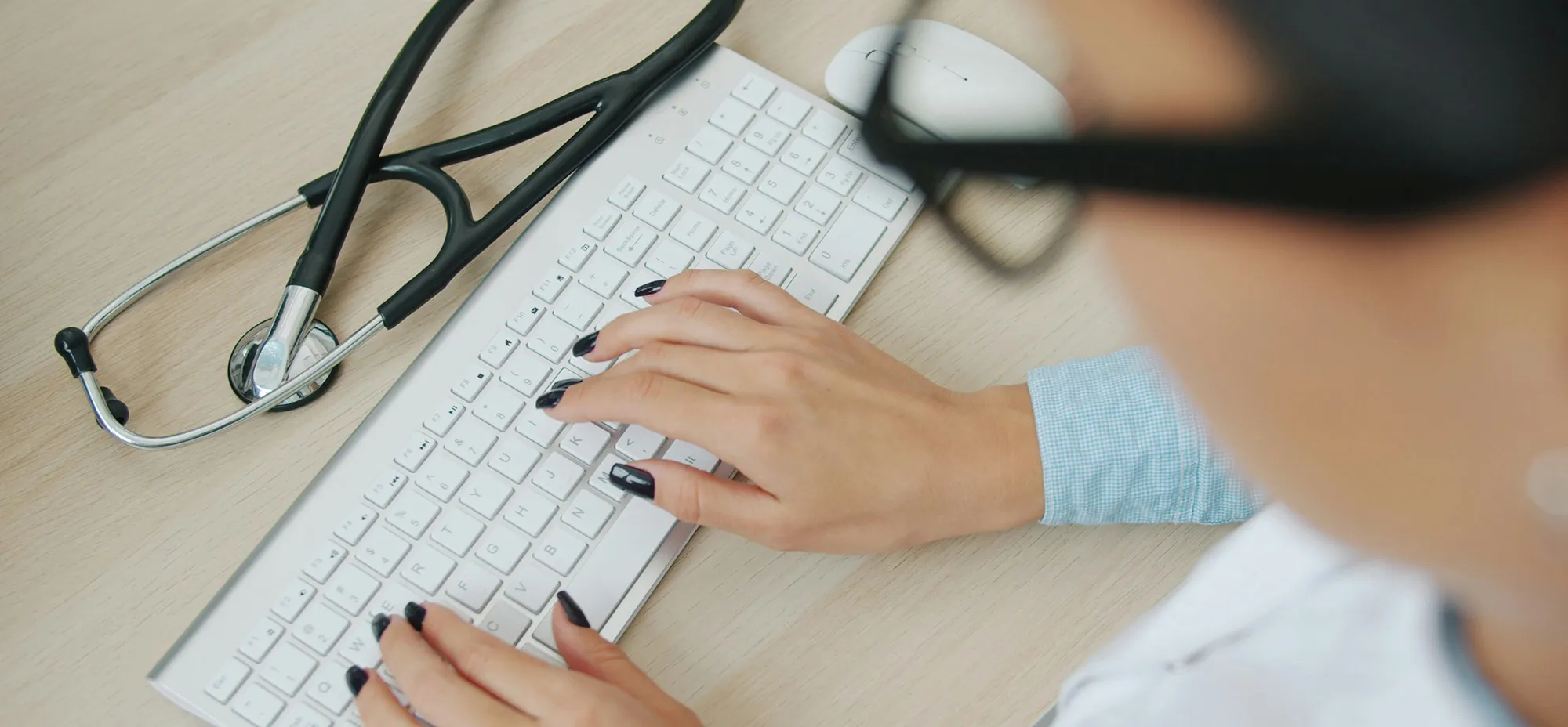 Overhead view of a person's hands with dark nail polish typing on a white keyboard, with a stethoscope and mouse resting on a wood desk.