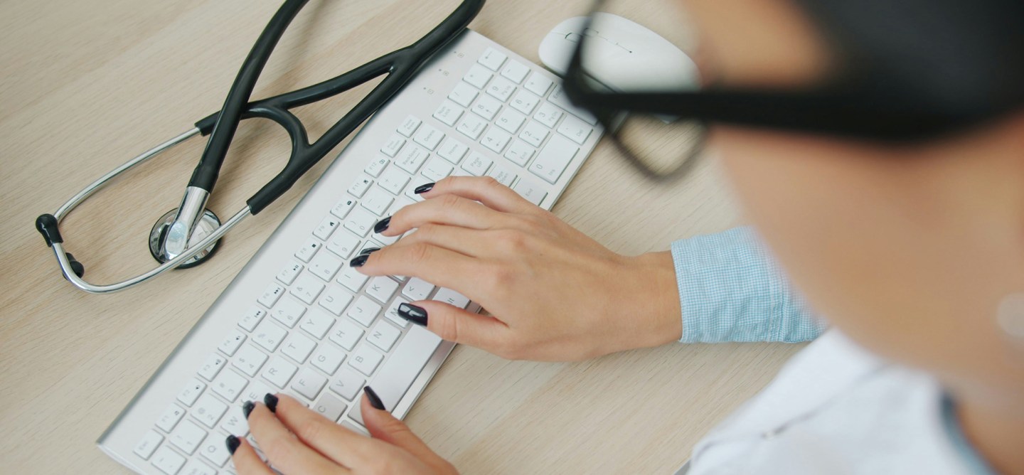 Overhead view of a person's hands with dark nail polish typing on a white keyboard, with a stethoscope and mouse resting on a wood desk.