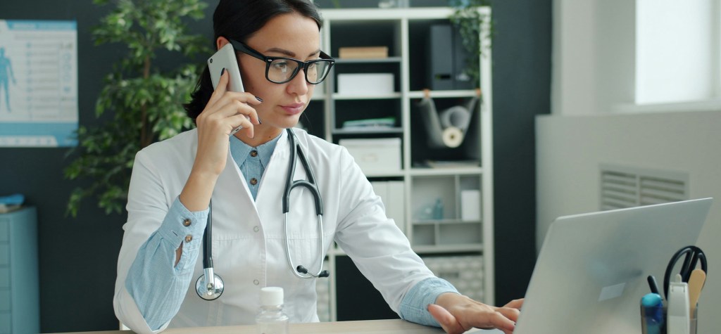 A doctor in a white coat and glasses talks on a mobile phone while working at a laptop in a bright medical office.