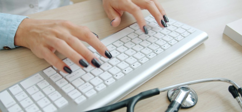 Close-up of hands with dark nail polish typing on a white keyboard beside a stethoscope on a wood desk.
