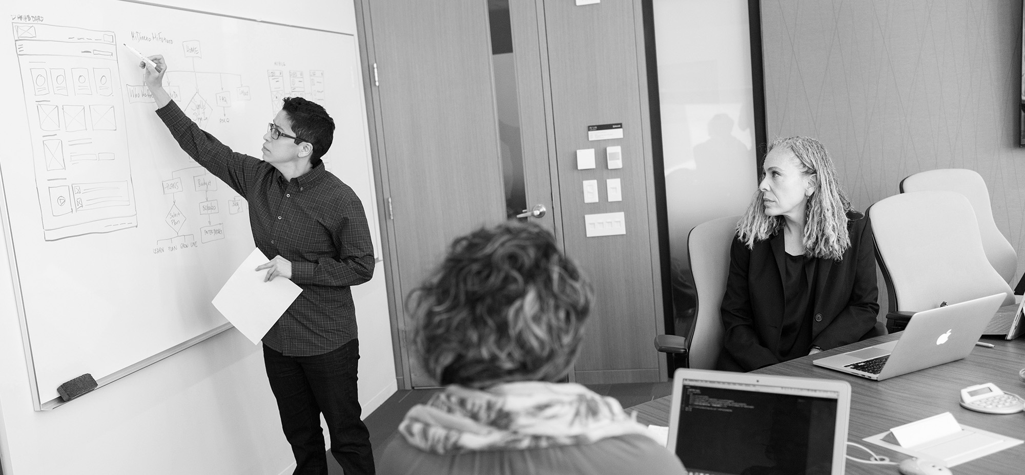 A team member sketches a website workflow on a whiteboard while two colleagues listen during a meeting.