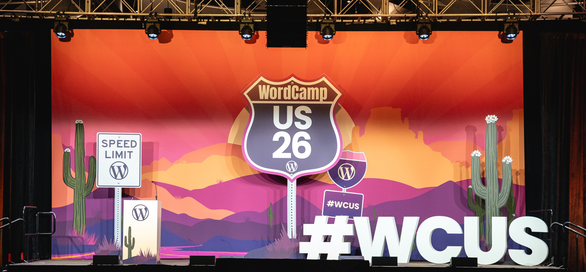 WordCamp US 2026 stage featuring a colourful desert-themed backdrop with cacti, WordPress logos, a large WCUS 26 road-sign graphic, and oversized #WCUS letters.