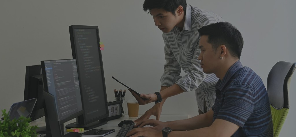 Two developers reviewing code together on multiple computer monitors at a workstation.
