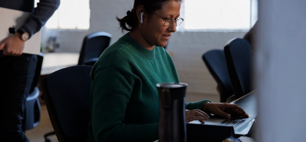 Woman wearing glasses and earbuds working on a laptop at a desk in an office.
