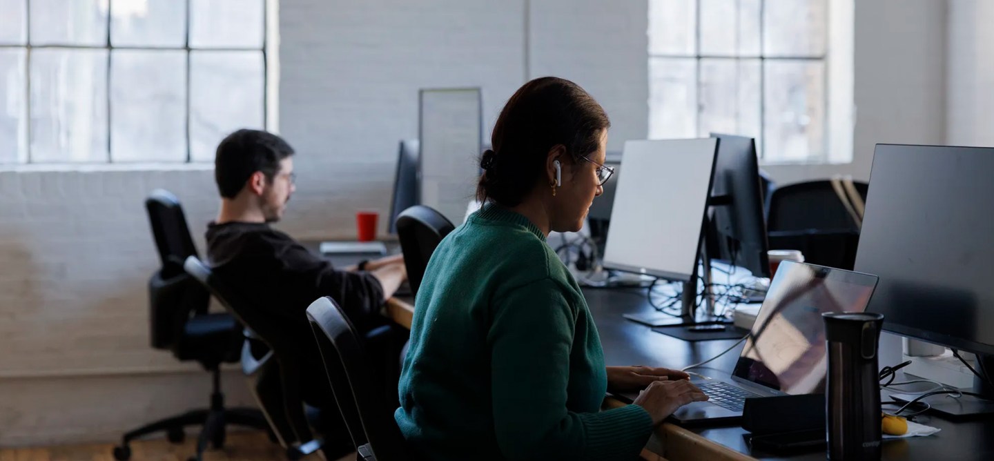 Woman wearing glasses and earbuds working on a laptop at a desk in an office.