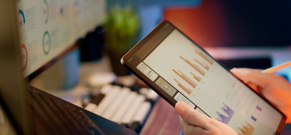 Close-up of a person holding a tablet displaying analytics dashboards and bar charts beside a computer monitor.